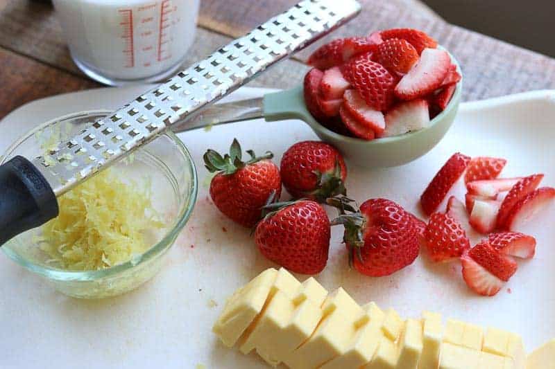 White cutting board with glass bowl full of lemon zest, zester, cut and whole strawberries, measuring cup with buttermilk and measuring cup with strawberries on a wooden board from Gourmet Done Skinny