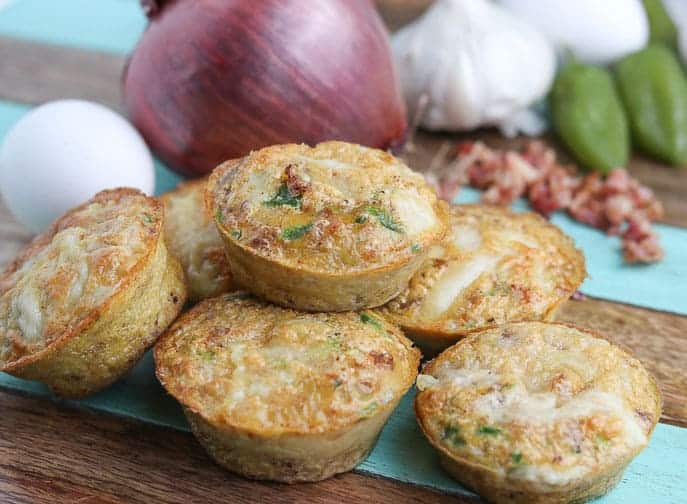 Delicious Chipotle Egg Cups ready to eat on a wooden board with red onion, egg, garlic, bacon and jalapeno in the background from Gourmet Done Skinny