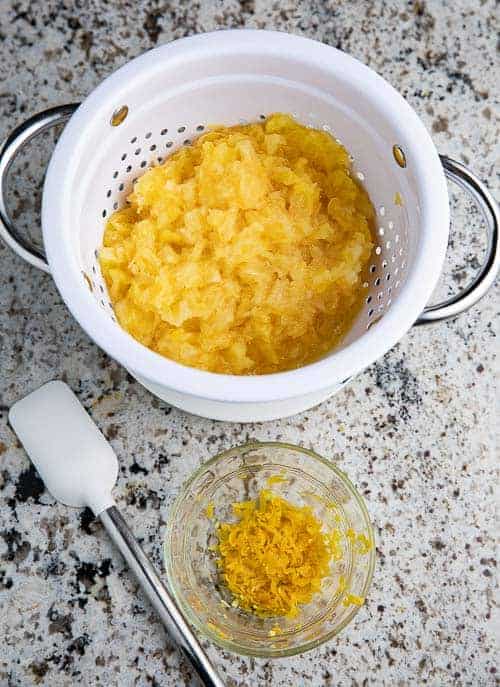 colander with crushed pineapple, orange zest in a glass bowl, white spatula on a granite countertop
