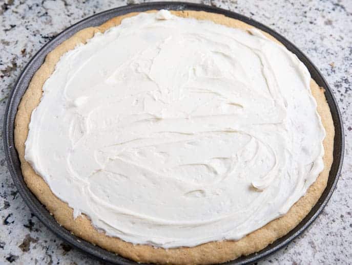 cookie crust covered with cream cheese spread on a pizza pan on a granite countertop