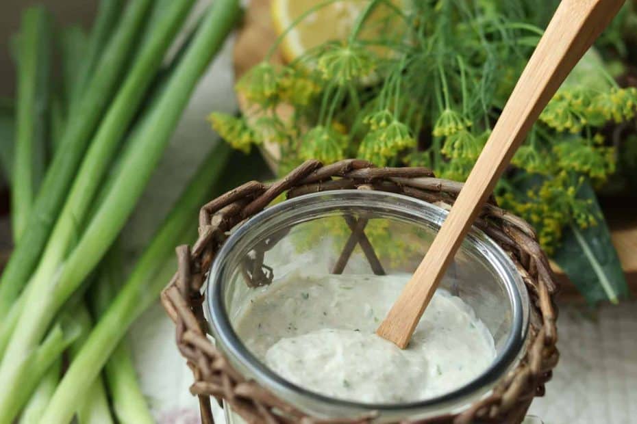 Healthy Garlic Ranch Dressing in a glass jar with wicker basket, green onions, dill and lemon on a fabric cloth