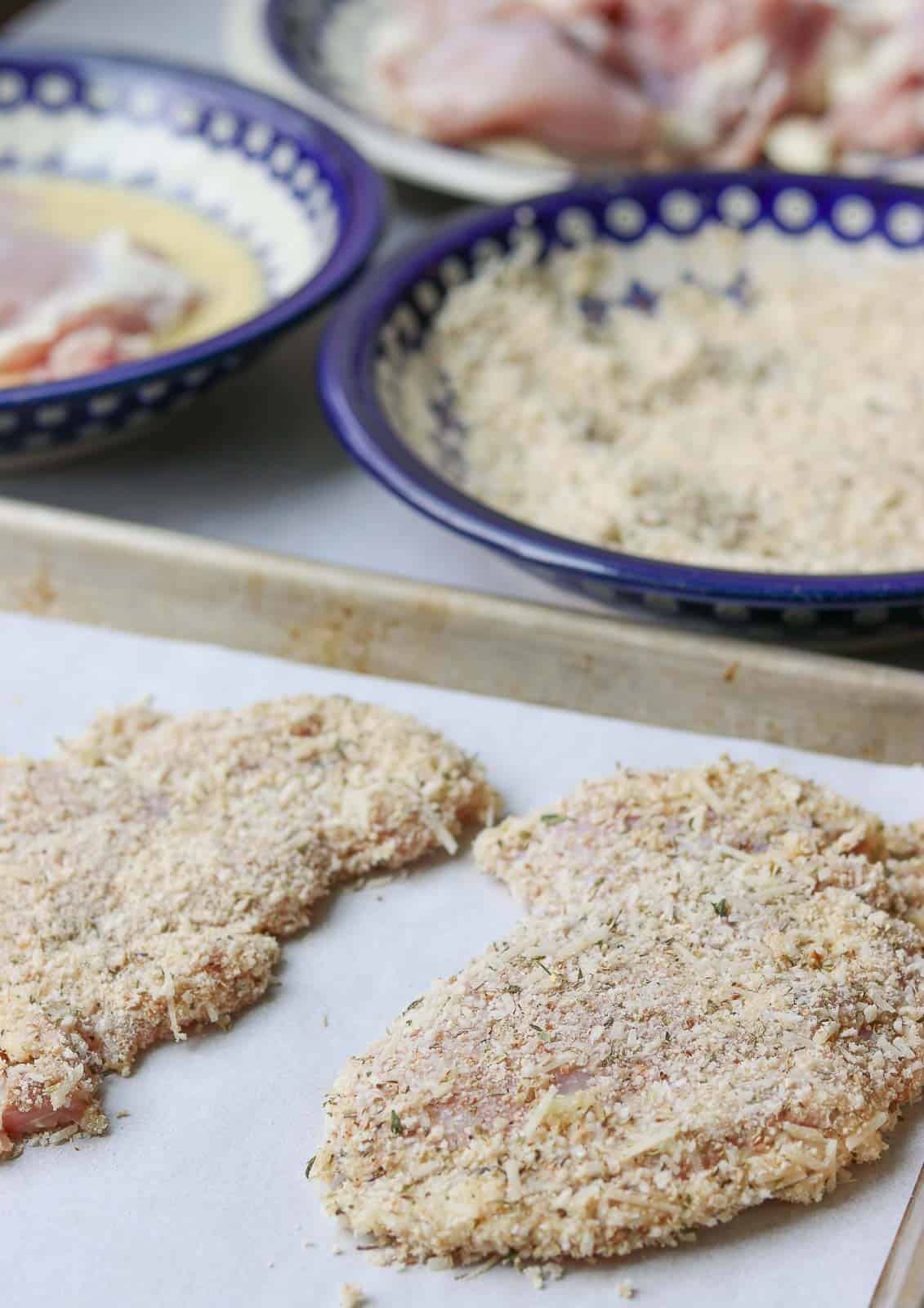 Parmesan Crusted Chicken raw on a baking sheet with egg mixture and crumb mixture in the background