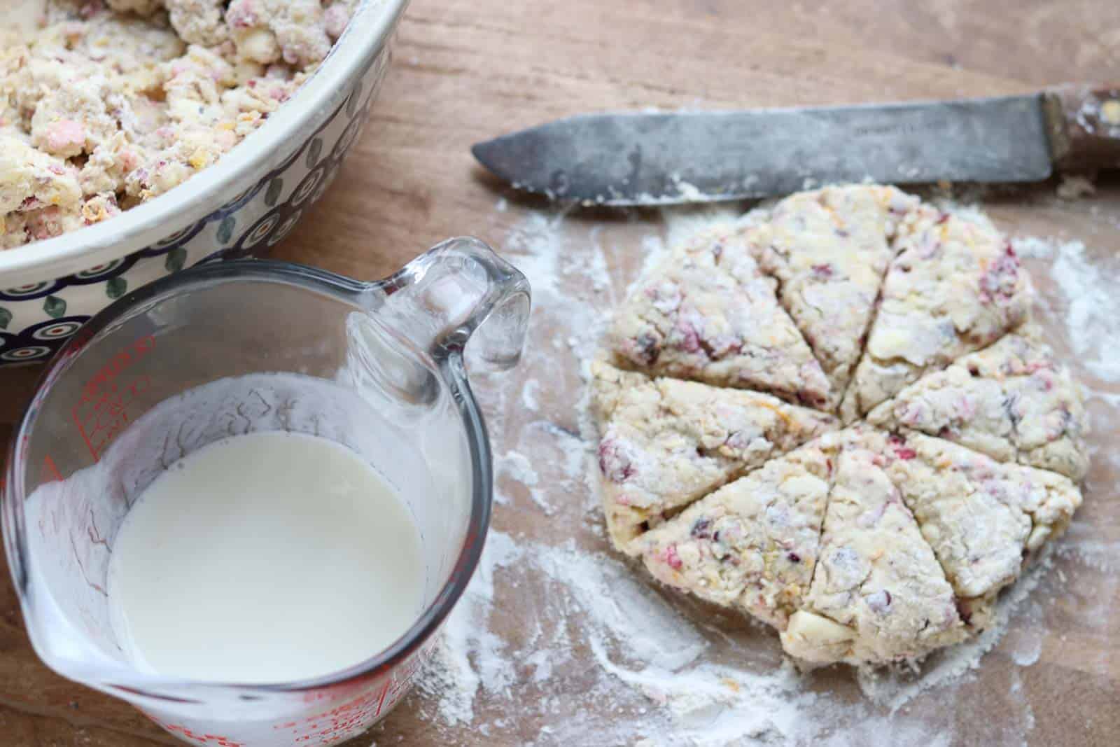 Scone dough cut into 8 pie shaped pieces, buttermilk in a liquid measuring cup and bowl of scone dough on a wooden board from Gourmet Done Skinny