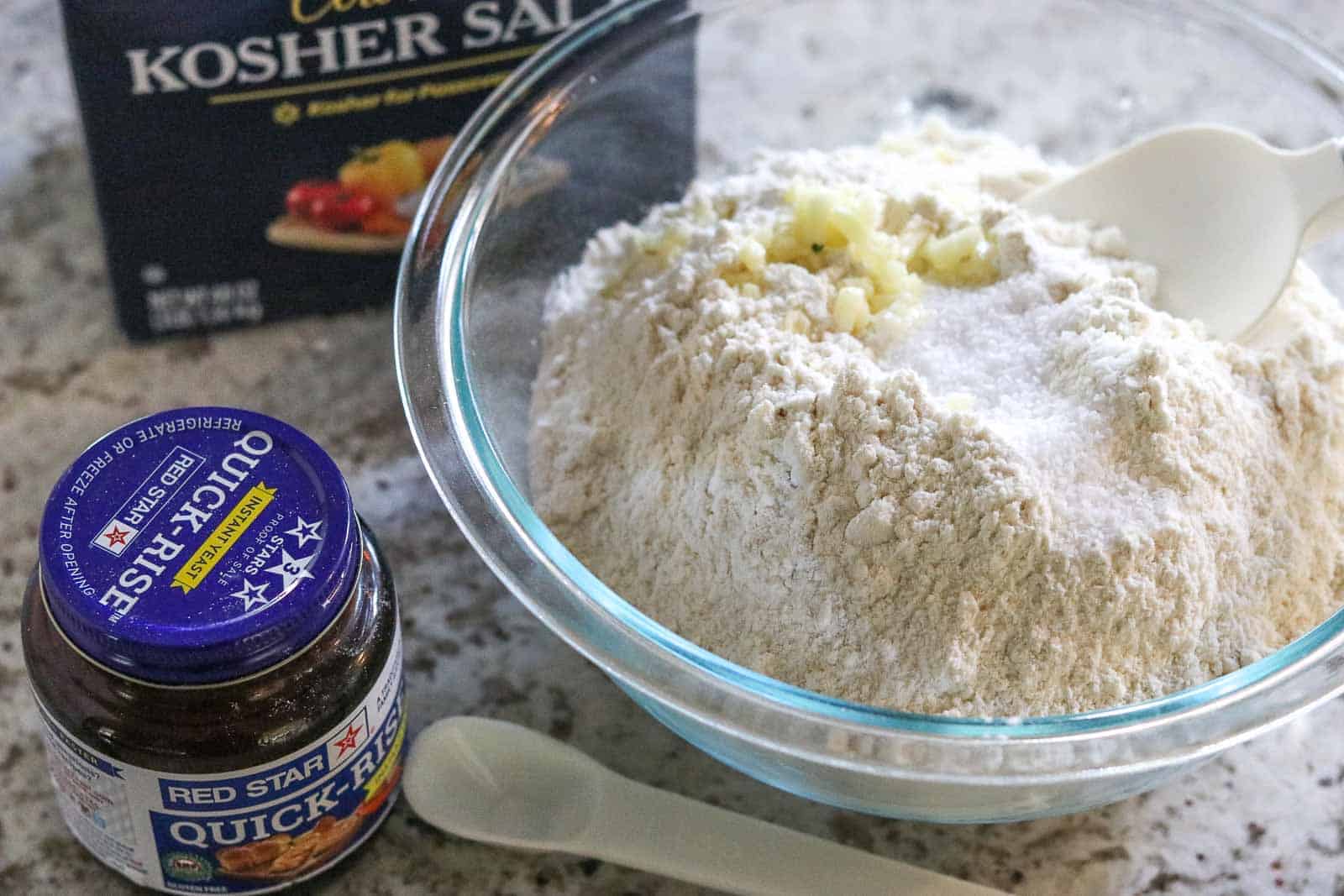 Glass bowl with flour, yeast, salt and minced garlic, with box of salt and jar of yeast in background from Gourmet Done Skinny
