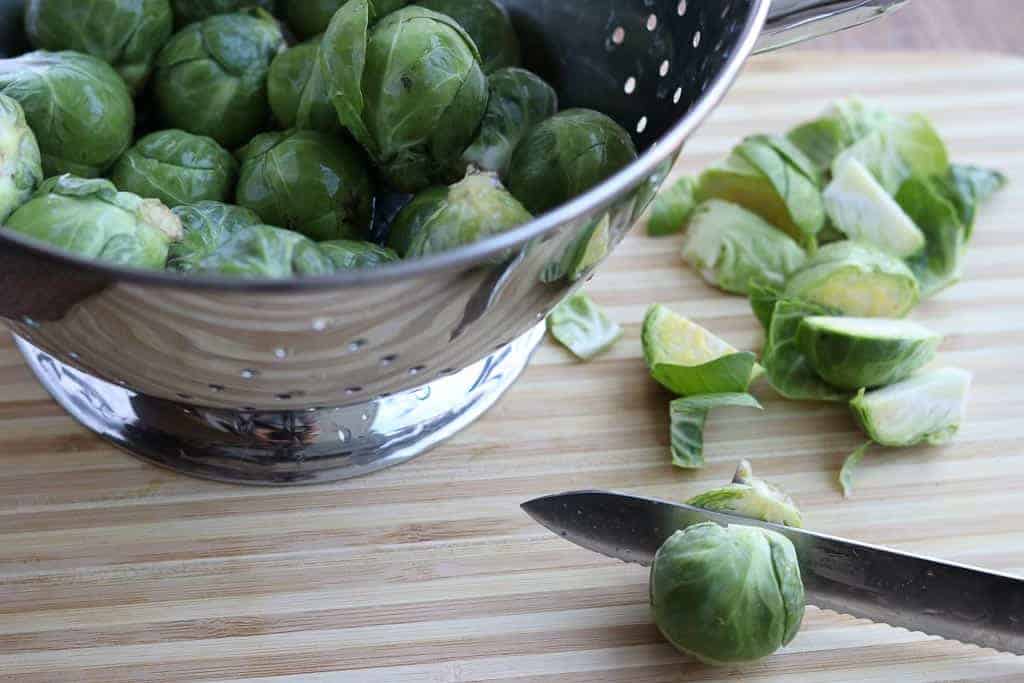 Metal colander with brussels sprouts on a wooden board with a brussels sprouts cut with a knife and cut brussels sprouts in the background from Gourmet Done Skinny