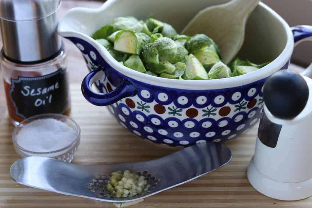 Sesame oil, salt, pepper, garlic press with garlic, Polish pottery bowl with brussels sprouts with a wooden spoon on a wooden board from Gourmet Done Skinny