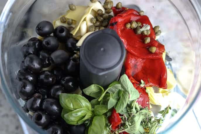Food processor mixer bowl with black olives, roasted red peppers, basil, thyme, capers and artichokes on a granite counter top from Gourmet Done Skinny