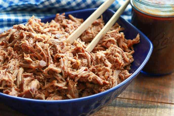 Easy healthy Instant Pot Pulled Pork in a blue bowl with a blue checked napkin and wooden tongs on a wooden board from Gourmet Done Skinny