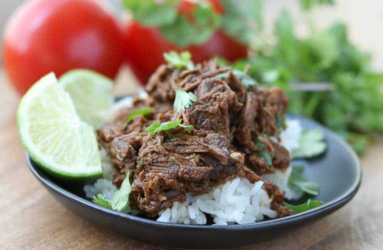 Easy Instant Pot Mexican Shredded Beef on a black plate with lime, cilantro and rice on a wooden board with tomatoes and cilantro in backgroundfrom Gourmet Done Skinny