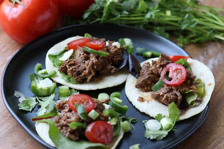 3 shredded beef tacos on corn tortillas on a black plate with tomatoes and cilantro in background on a wooden board from Gourmet Done Skinny