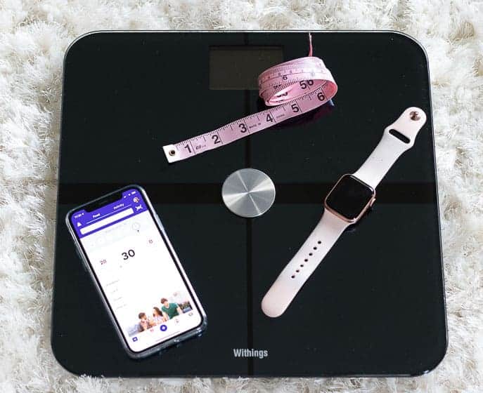 Black withings scale with pink tape measure, iphone with Weight Watcher Ap, Apple Watch on a white carpet from Gourmet Done Skinny