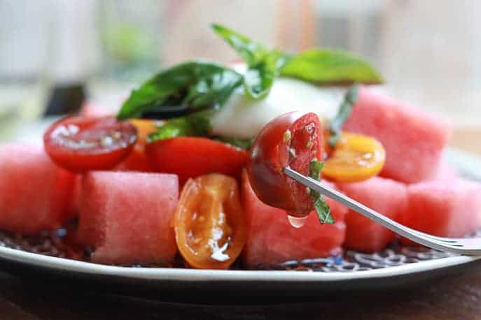 Tomatoes and watermelon pieces on plate with burrata, mint and basil shreddings, wine glasses blurred in background on a wooden board from Gourmet Done Skinny