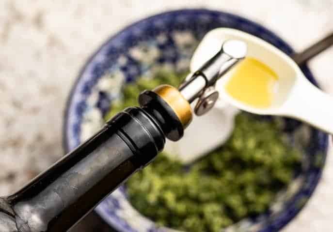 Bottle of olive oil being poured into a tablespoon over pesto
