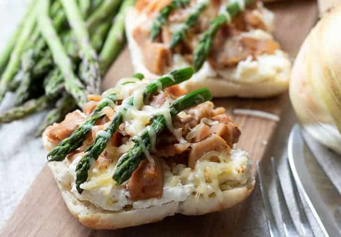 Wooden board with 2 sandwich halves topped with caramelized onion filling, topped with grilled chicken, asparagus and gruyere cheese