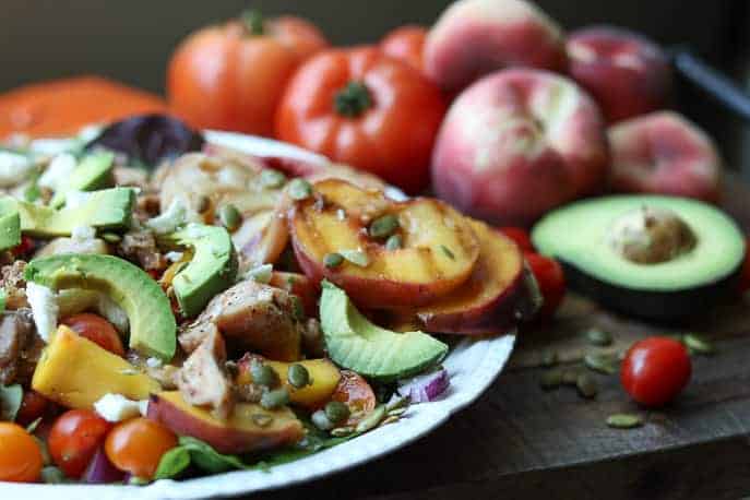 Summer Chicken Salad with fresh tomatoes, grilled peaches, avocados, pumpkin seeds on a white plate with tomatoes, peaches, avocados in the background