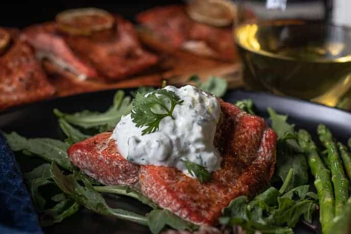 Cedar plank salmon on top of greens with asparagus on a black plate with salmon on Cedar planks in background and a glass of white wine from Gourmet Done Skinny