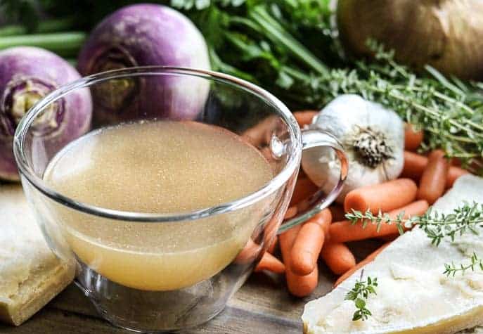 Glass cup with Parmesan Chicken broth with turnips, carrots, garlic, herbs and parmesan rinds on a wooden board from Gourmet Done Skinny