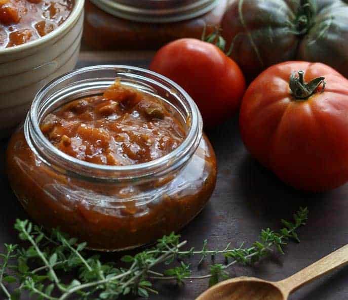 Tomato Bacon Jam in a glass jar with tomatoes, thyme and wooden spoon by Gourmet Done Skinny.