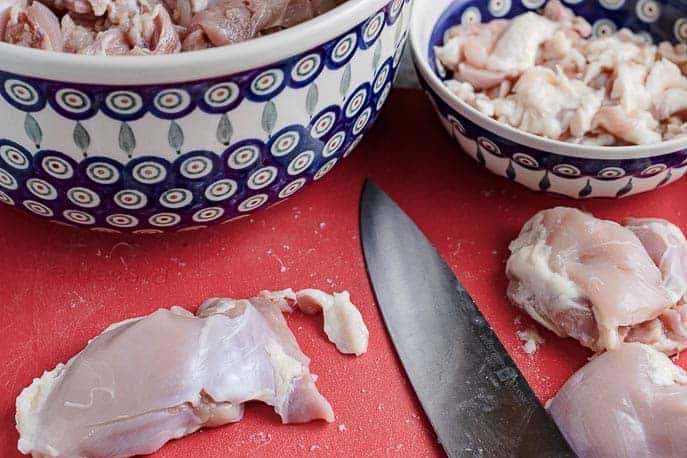 Chicken thigh on a cutting board with knife, chicken fat in a small bowl, cut chicken in a big bowl from Gourmet Done Skinny