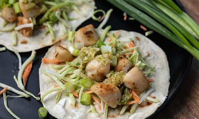 Pan Seared Scallop Tacos on a black plate with green onions in background