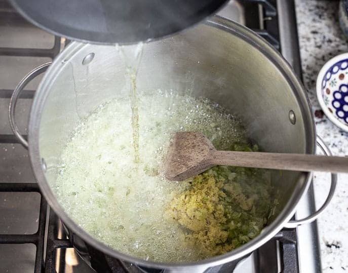 pot with oil pouring out into a large pot with green onion mixture, wooden spoon and mixture sizzling and steaming
