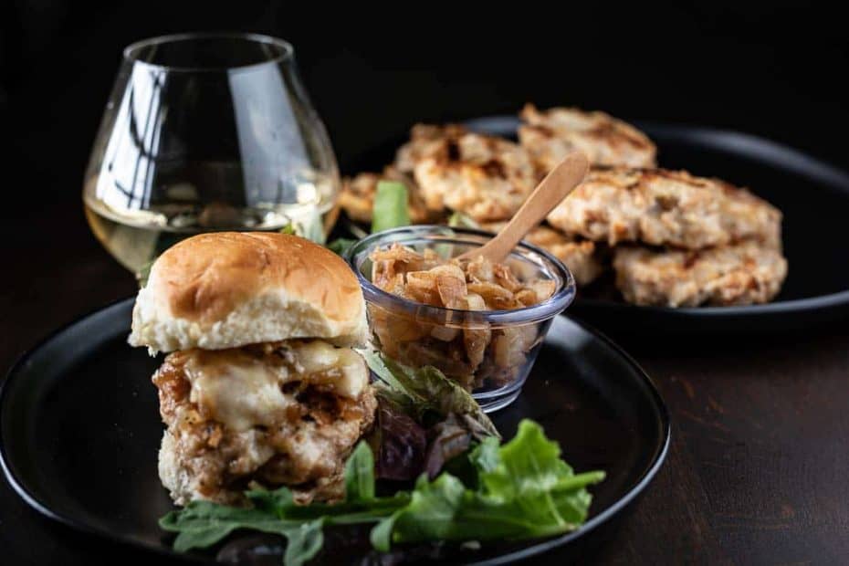 Healthy chicken burger with caramelized onions on a black plate with wine and more patties in the background