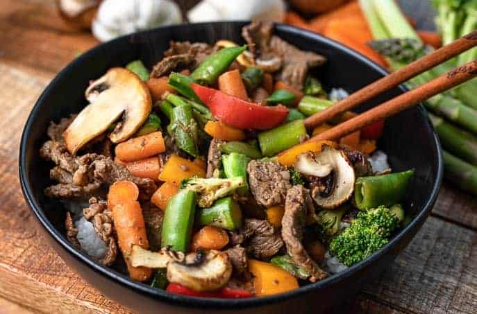 Clean Out Your Refrigerator Stir Fry in a black bowl on a wooden board with chop sticks and vegetables in the background