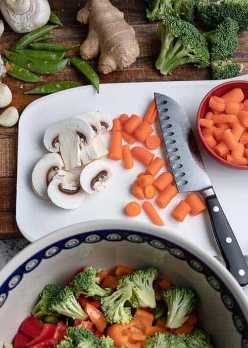 Cutting board with cut carrots and mushrooms, vegetables in background and bowl of vegetables in foreground