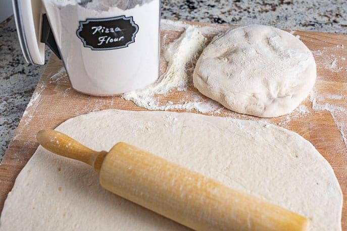 pizza dough rolled out on a wooden board with flour and ball of dough in the background