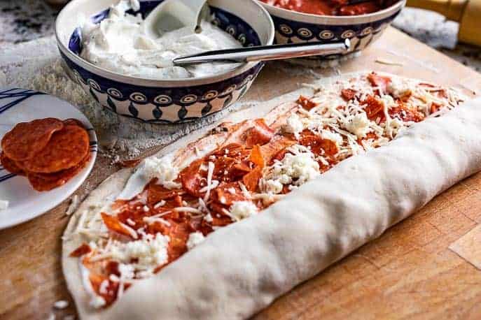 Pasta per pizza con formaggio, peperoni parzialmente arrotolati su una tavola di legno con peperoni, salsa sullo sfondo