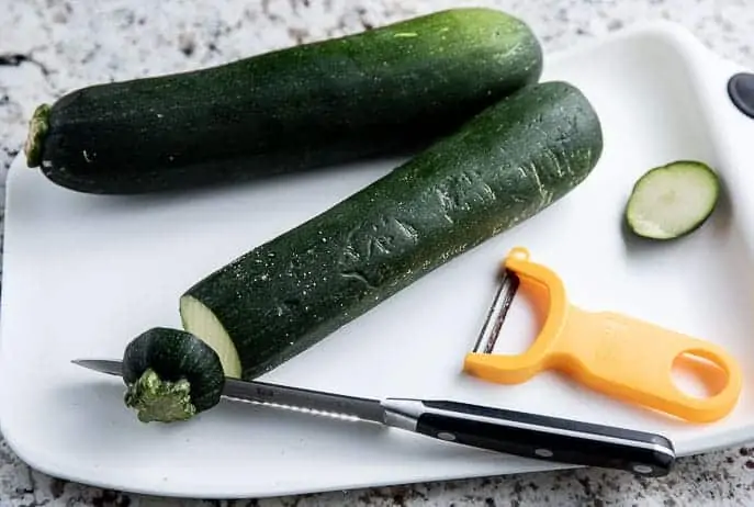 2 zucchini on a white cutting board with knife and y peeler
