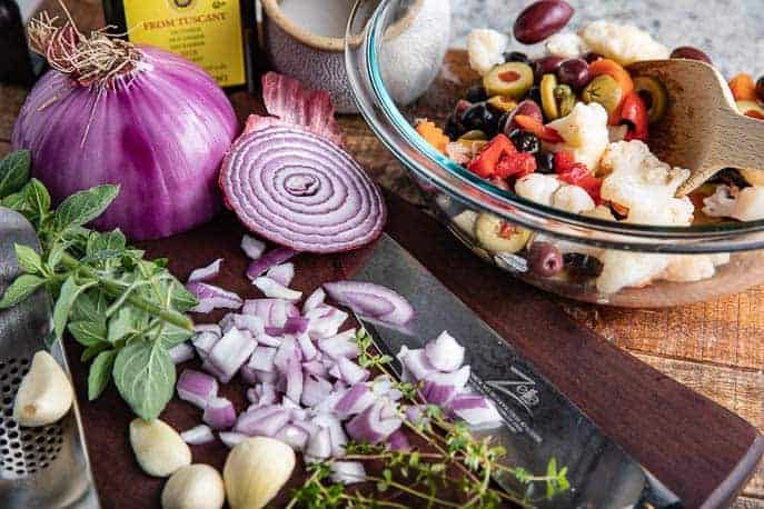Ingredients for Muffuletta salad - onions, garlic, thyme, oregano, glass bowl with olives, italian vegetables, kosher salt, olive oil on dark and light wooden boards