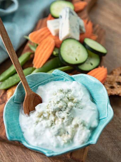 Blue bowl with blue cheese dressing, wooden spoon, pretzels, carrots, blue cheese wedge, on a wooden board