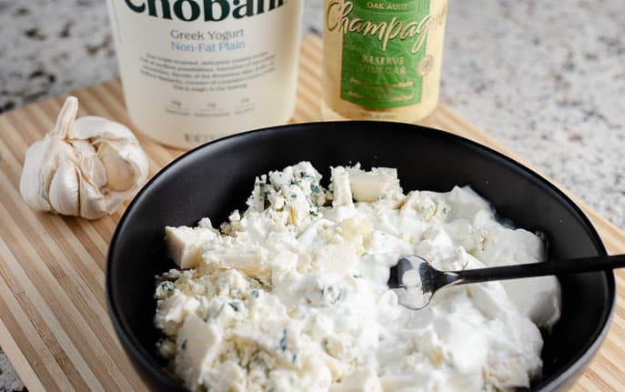 blue cheese and yogurt in a black bowl with garlic cloves, yogurt container and champagne vinegar on a wooden cutting board