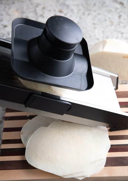 Thinly sliced jicama on a striped cutting board with a mandoline on a granite counter top