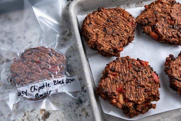 Spicy Chipotle Black Bean Burgers on a baking sheet frozen, vacuum sealed bag with one burger next to pan