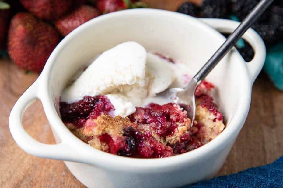 small white dish with handles with mixed berry cobbler and a black spoon, strawberries and blackberries in background