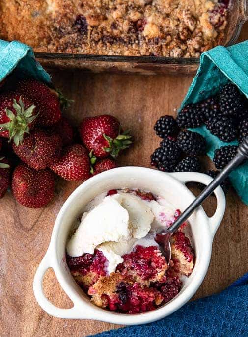 cobbler and ice cream with black spoon on a wooden board with blue napkin, fresh strawberries and blackberries and cobbler in a baking dish in back