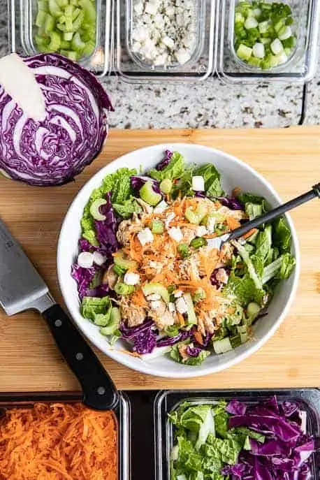 white bowl with buffalo chicken, cabbage, celery, blue cheese, green onions, cabbage and carrots in plastic containers