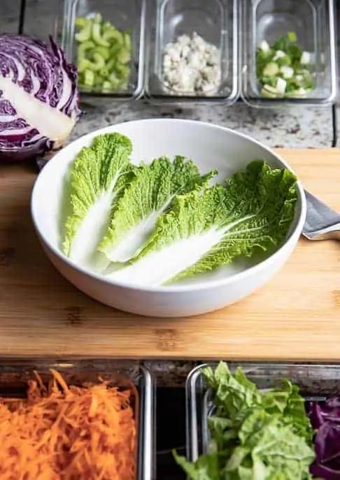 white bowl with cabbage leaves, celery, blue cheese, green onions, cabbage and carrots in plastic containers