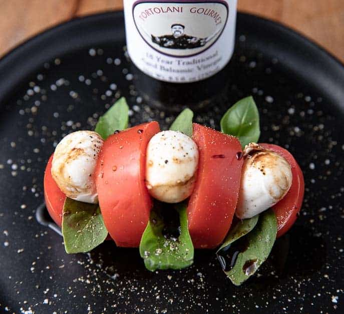 tomato sliced hasselback style on a black plate with basil, mozzarella and balsamic vinegar