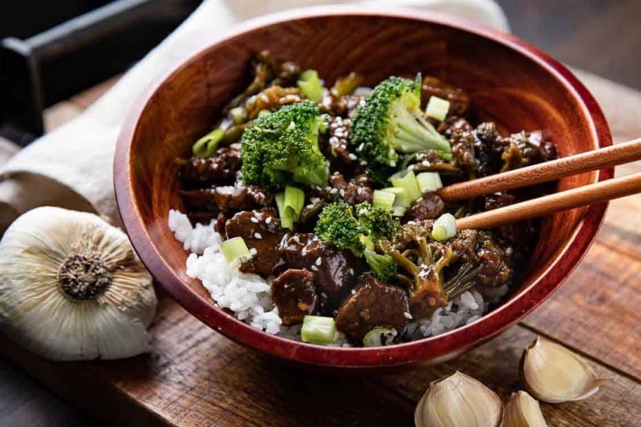 Mongolian beef and broccoli in a wooden bowl with rice and chopsticks on a wooden board with garlic cloves around