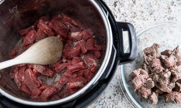 flank steak in Instant Pot with wooden spoon next to glass bowl with browned flank steak