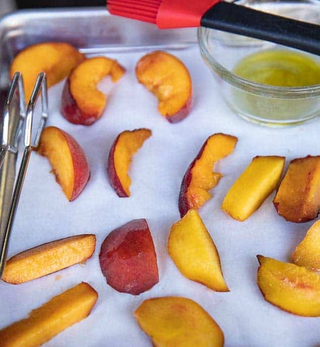 peach slices on a baking sheet with parchment, olive oil in glass bowl with pastry brush and tongs
