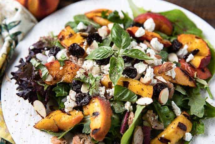 Grilled peach salad - Salad greens with peaches, chicken, goat cheese, basil, dried cherries on a white plate with peach and colored napkin in background