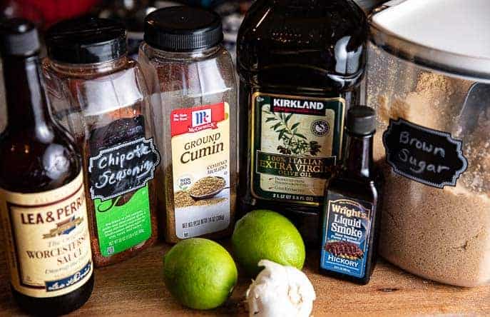 Ingredients for fajita marinade on a wooden board - worcestershire sauce, chipotle seasoning, cumin, oil, liquid smoke, brown sugar, 2 limes and head of garlic