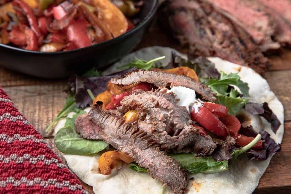 Grilled Steak on a tortilla with greens on a wooden board with vegetables and flank steak and napkin in the background