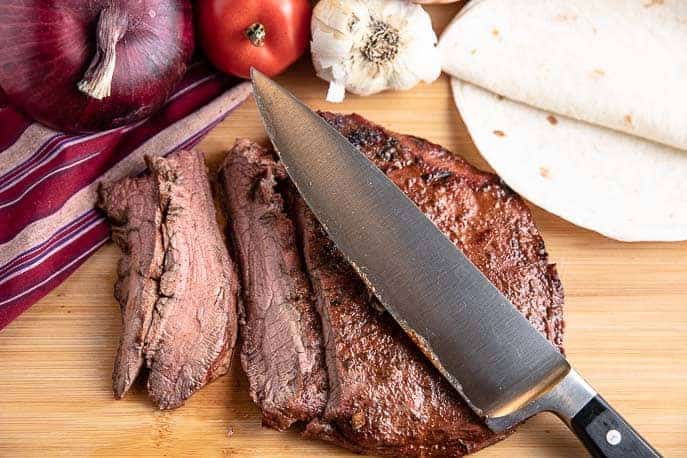 grilled flank steak on a cutting board with knife, onion, tomato, peppers, garlic and tortillas in background