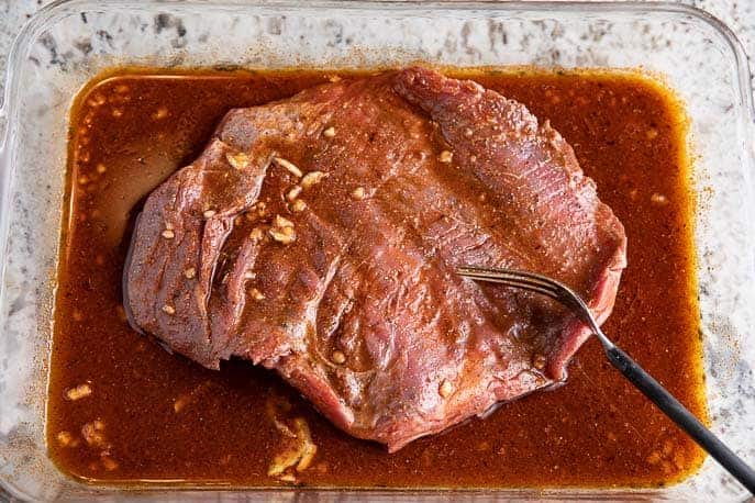 Flank steak marinating in a glass dish with black fork