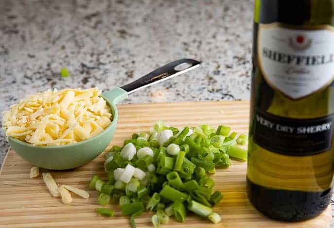 white cheddar cheese in a measuring cup and chopped green onions on a cutting board with a bottle of sherry